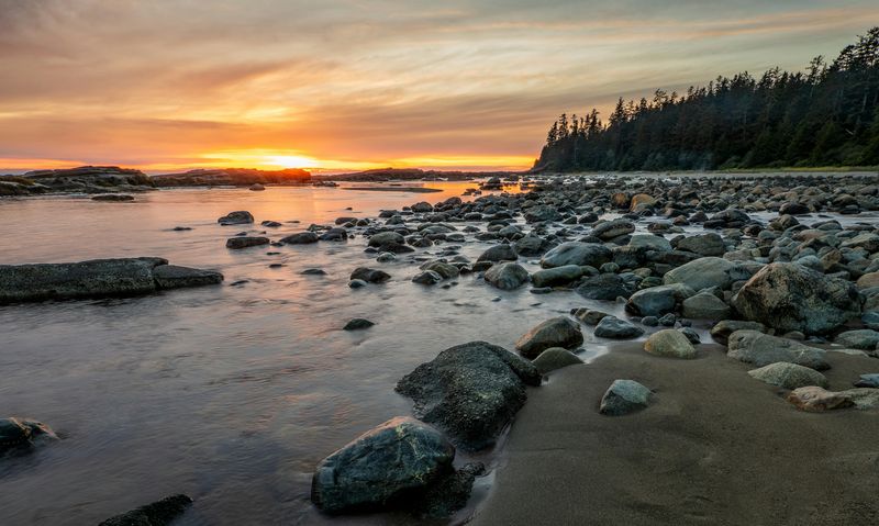 A scenic landscape featuring a rocky shoreline at sunset, with smooth water reflecting the orange and yellow hues of the sky and trees in the background.