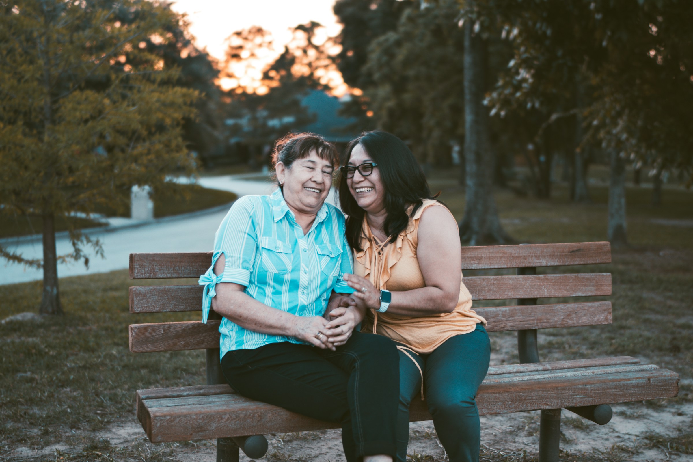 Two women sitting on a bench, laughing and enjoying each other's company in a park during sunset.